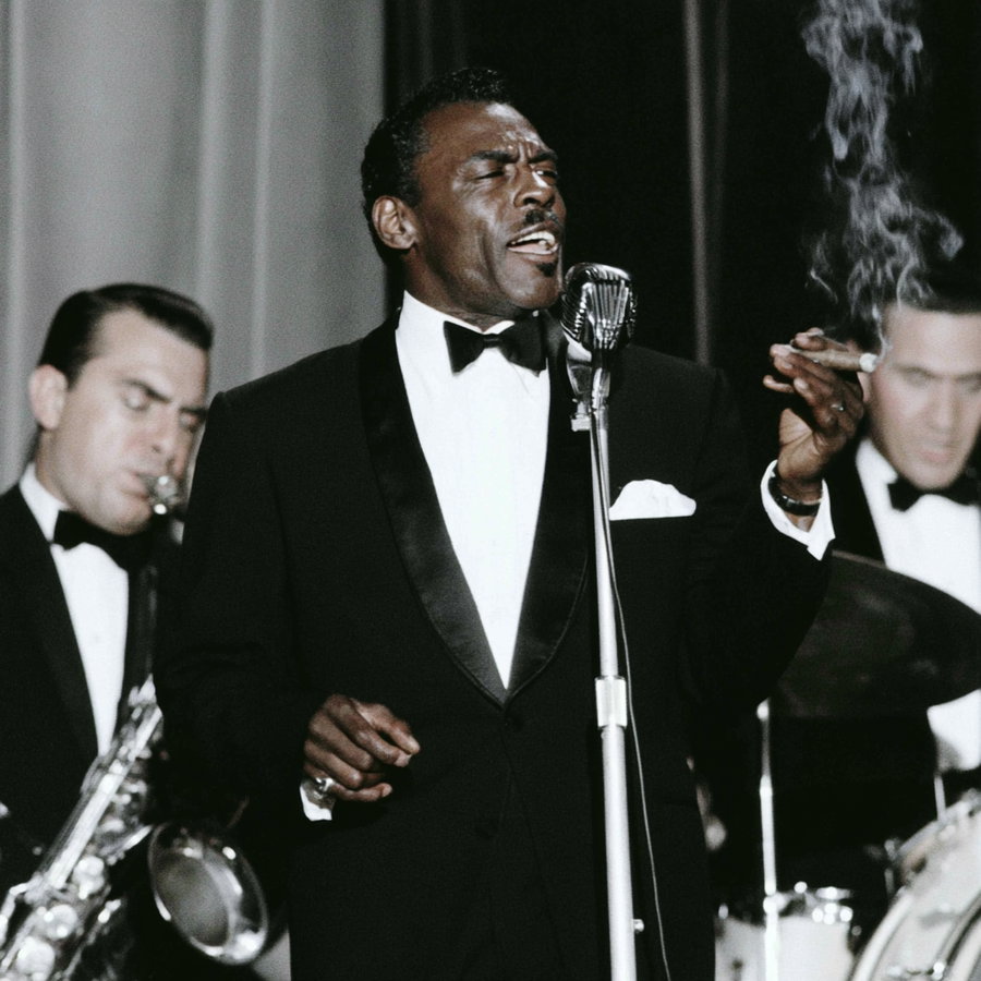 Black male singer in tuxedo holding a cigar and singing into a microphone, with musicians playing saxophone and drums in the background
