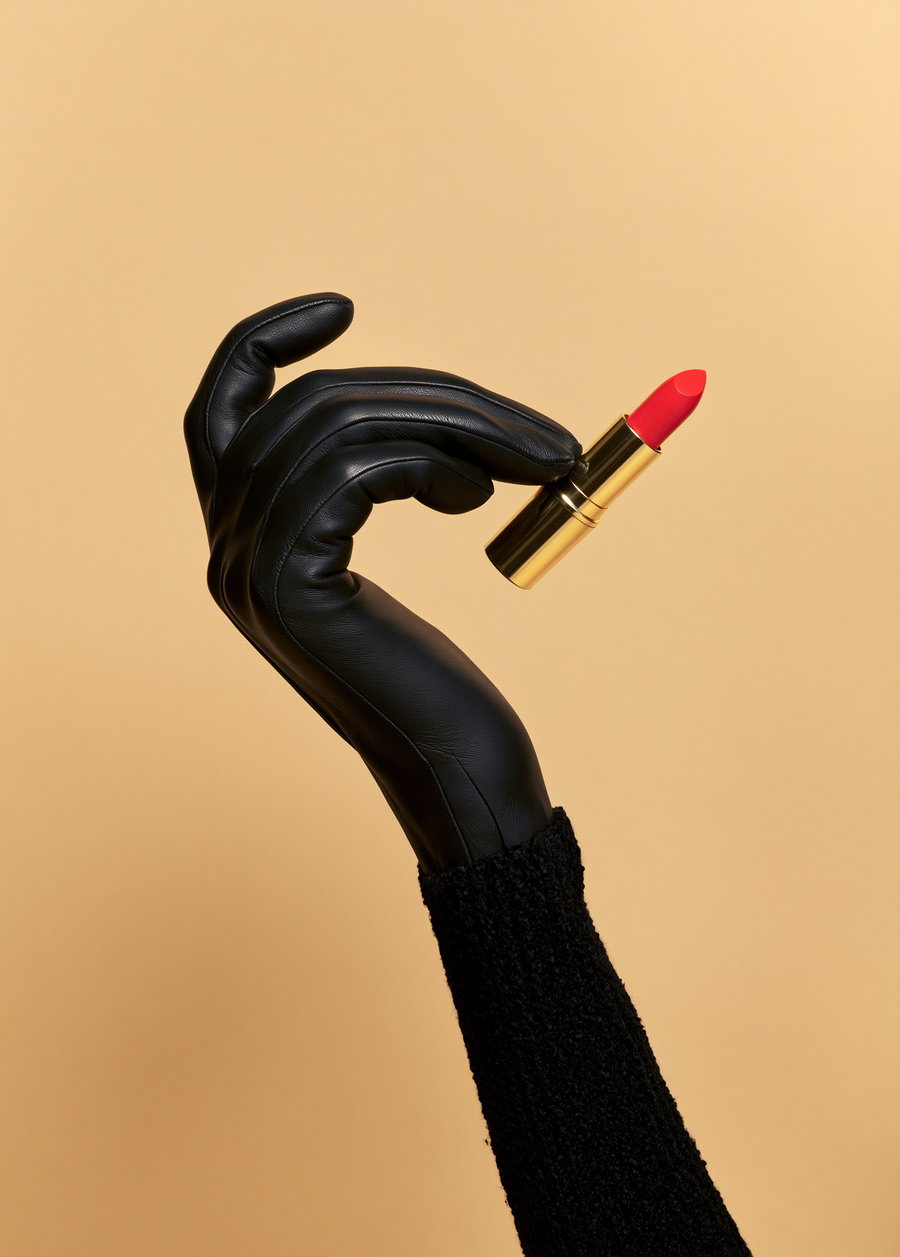 A close-up of a hand wearing a black leather glove and black textured sleeve holding a gold and red lipstick against a beige background