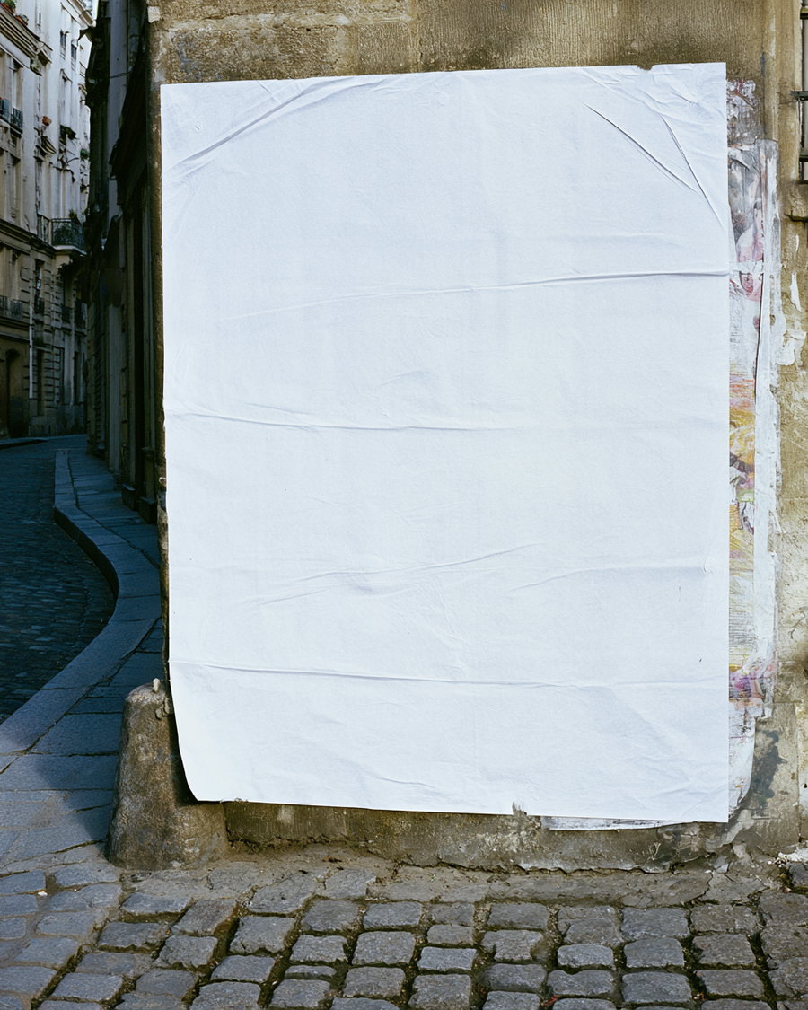 a large blank white paper poster attached to a weathered urban wall on a cobblestone street corner with old buildings in the background