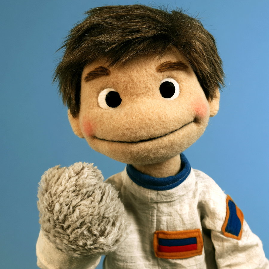 Close-up image of a puppet with brown hair, large black eyes, and a subtle smile, dressed in a white costume with blue, orange, and red patches against a blue background