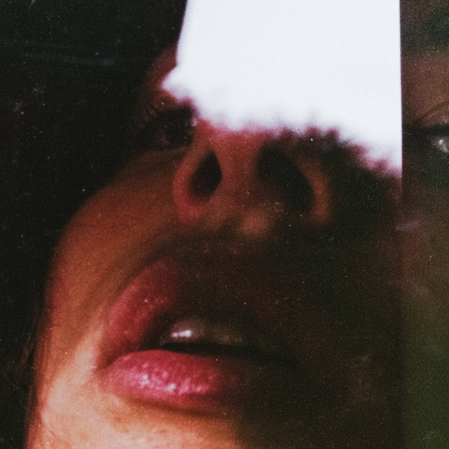 A close-up of a person's face partially obscured by a reflective glass with shadows and highlights emphasizing lips, nose, and eyes.