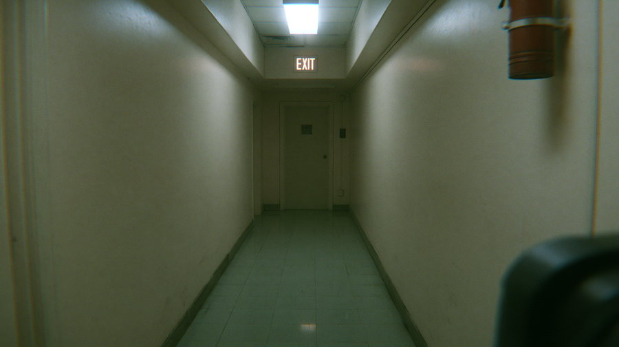 A narrow, dimly lit corridor with pale walls and a tiled floor leading to a closed door under an illuminated exit sign