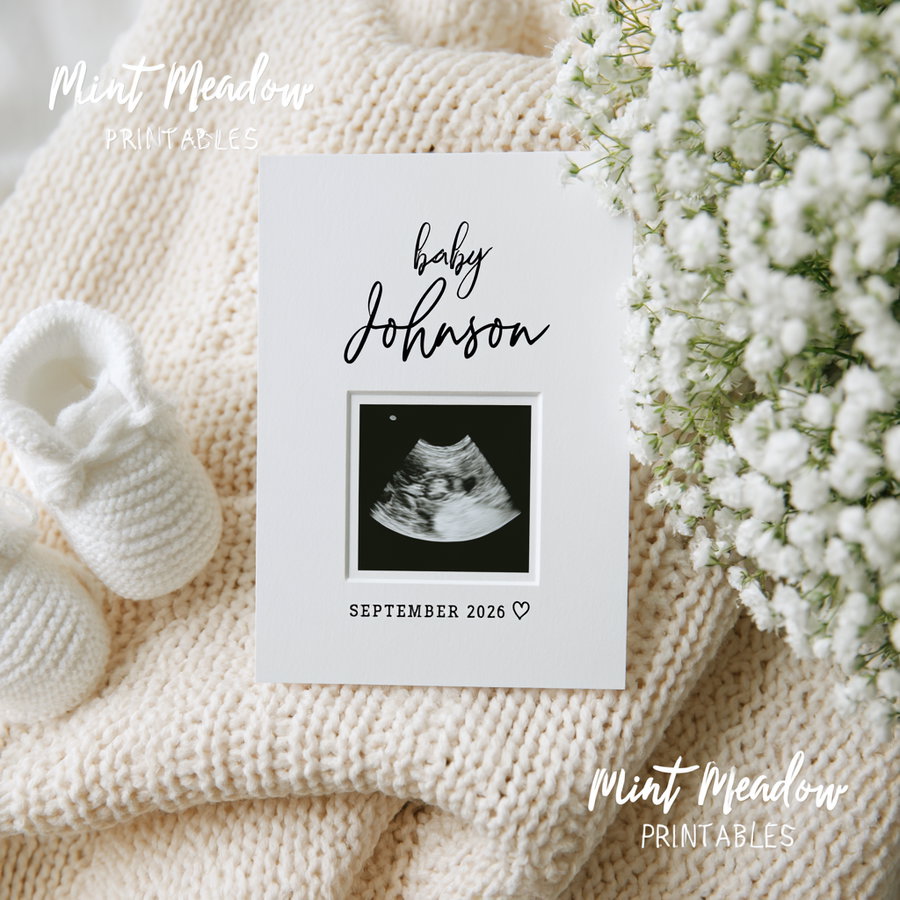 A baby ultrasound photo in a white frame with the text baby Johnson and September 2026, placed on a cream knitted blanket alongside white knitted baby booties and white flowers