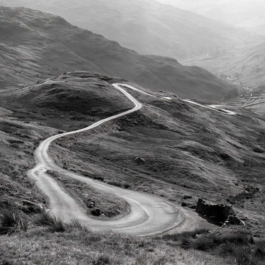 Black and white image of a winding road traversing rolling hills with textured grassy landscape