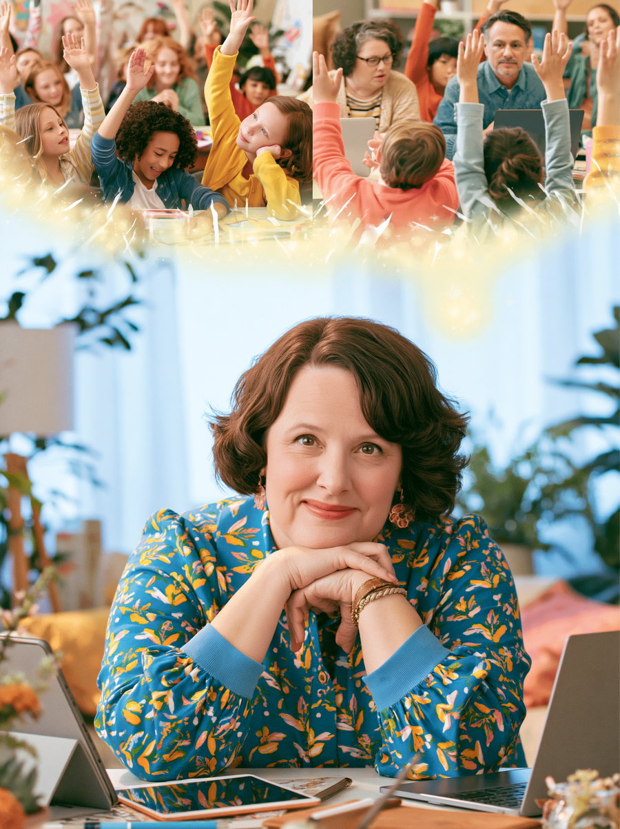 A woman with short curly hair in a blue floral blouse smiles gently with her hands under her chin, sitting at a desk with laptops and tablets in a bright, cozy room. Above her, children and adults are actively raising their hands, engaging in classroom activities.
