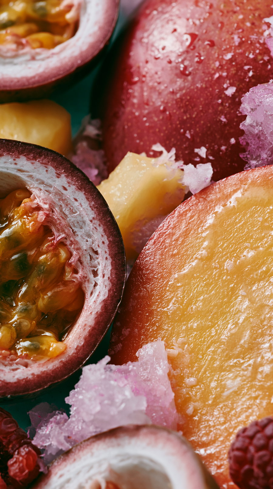 Close-up image showing sliced passion fruit, mango, pineapple, red apple, raspberries, and crushed ice