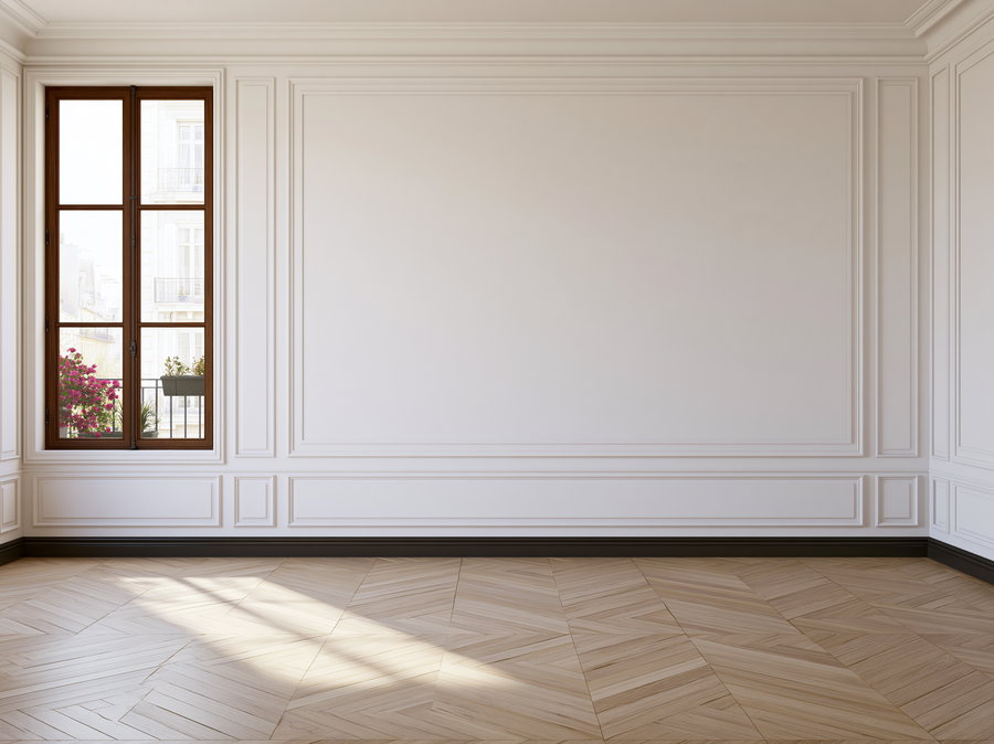 Empty room interior with white walls featuring decorative molding, wooden floor, and a window with a view of flowers outside