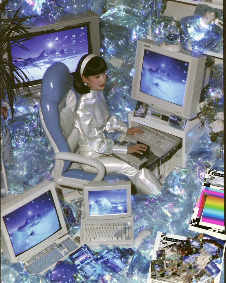 A woman dressed in shiny silver clothing seated in a blue and white chair operating a vintage computer surrounded by multiple old computer monitors showing a surreal digital landscape with icy mountains and a glowing star, all placed within a room filled with iridescent, crumpled foil-like material and various scattered printed images.