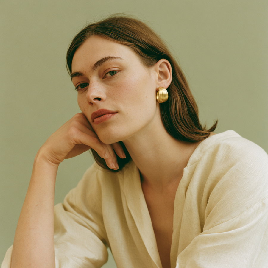 Close-up of a woman with light skin, green eyes, and brown hair wearing a cream-colored top and large gold earrings against a muted green background