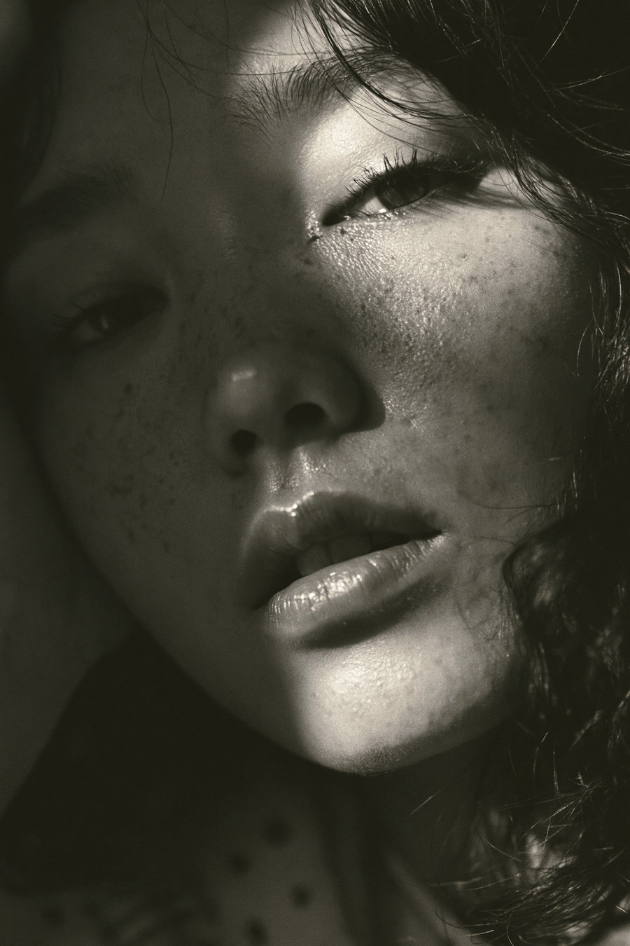 A close-up grayscale portrait of a woman with freckles, partially shadowed with soft light highlighting her lips, nose, and one eye