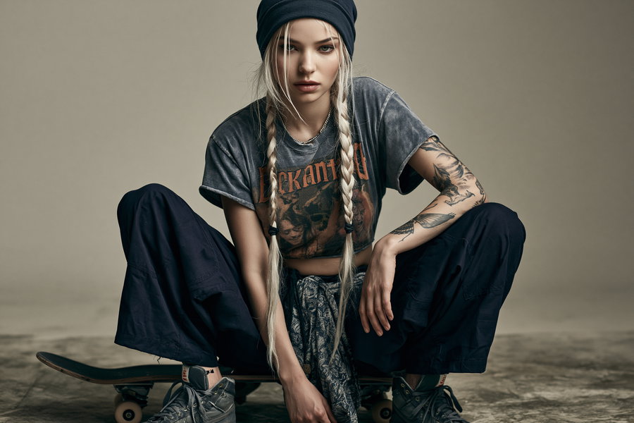 A young woman with long braided blonde hair, wearing a black beanie, cropped graphic t-shirt, black pants, and sneakers, sitting on a skateboard against a neutral background