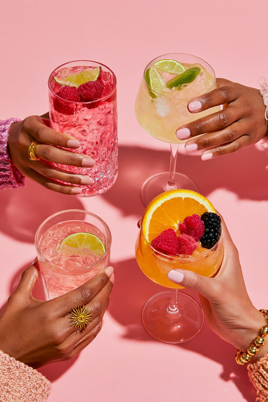 Four hands holding different glasses filled with colorful cocktails garnished with lime, orange slices, raspberries, and blackberries on a solid pink surface
