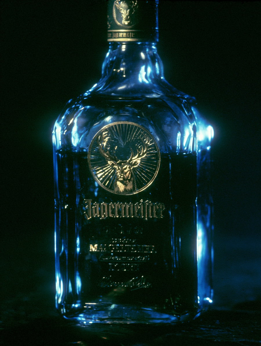 A close-up image of a Jägermeister bottle with a golden stag emblem and dark liquid inside, illuminated by blue lighting against a black background.
