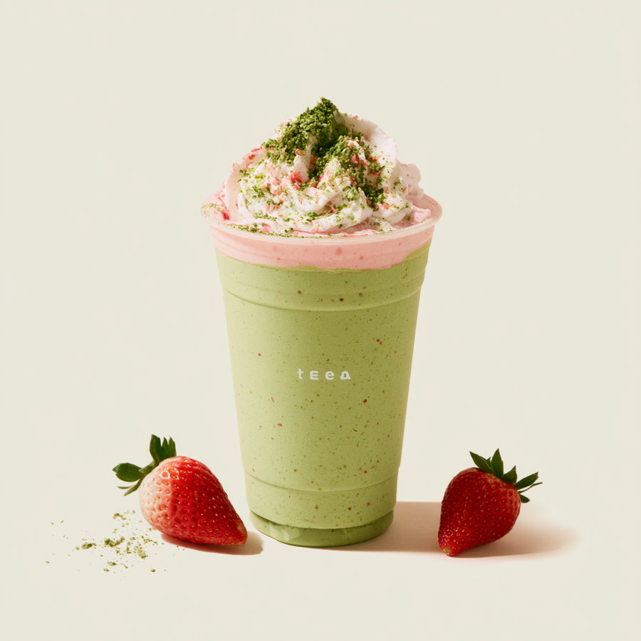 A close-up image of a green matcha smoothie or drink with pink whipped cream on top and green powder sprinkled, placed between two red strawberries on a light background.