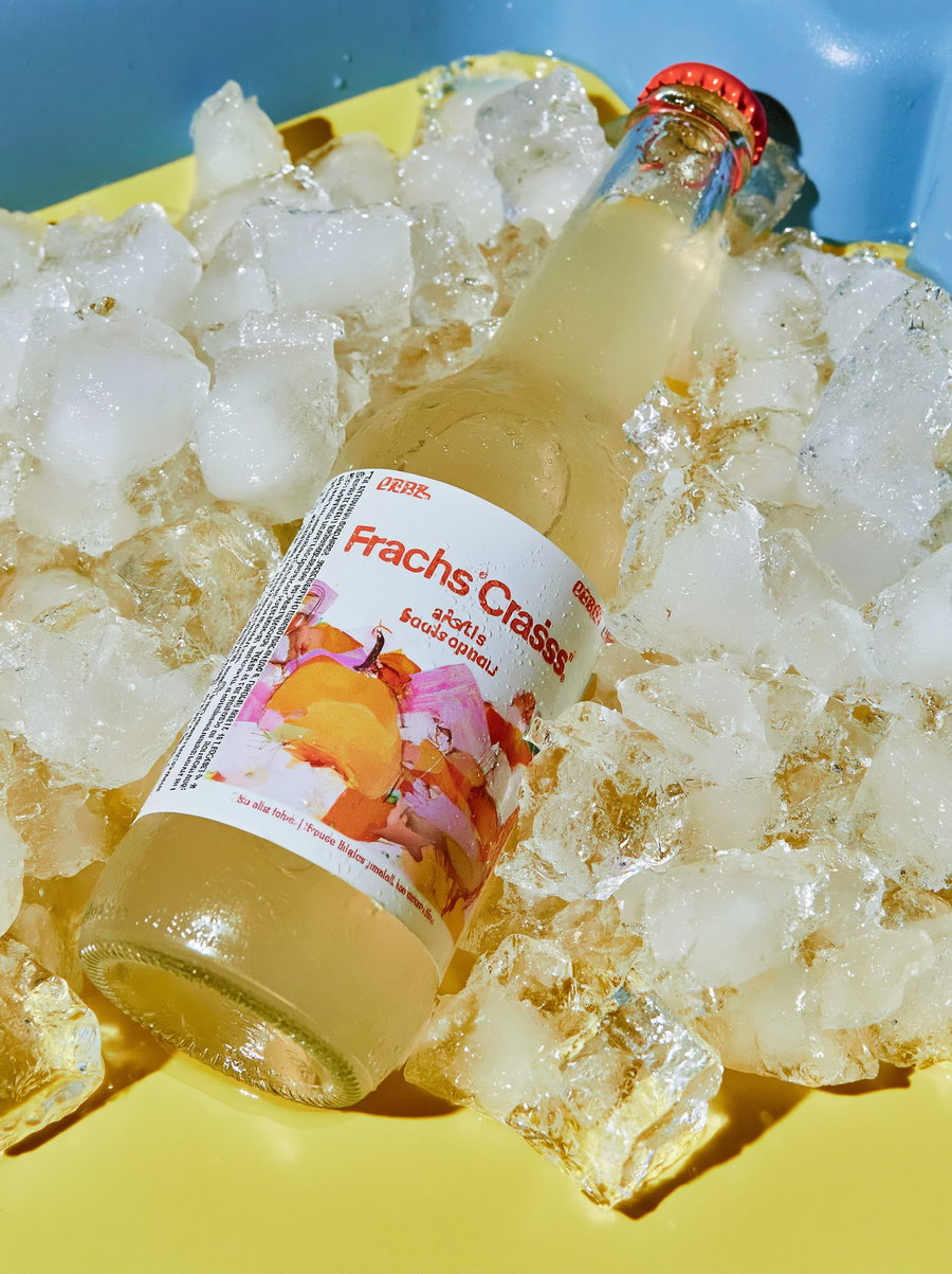 A glass bottle with an abstract painted label lying on ice cubes in a yellow container with some blue visible