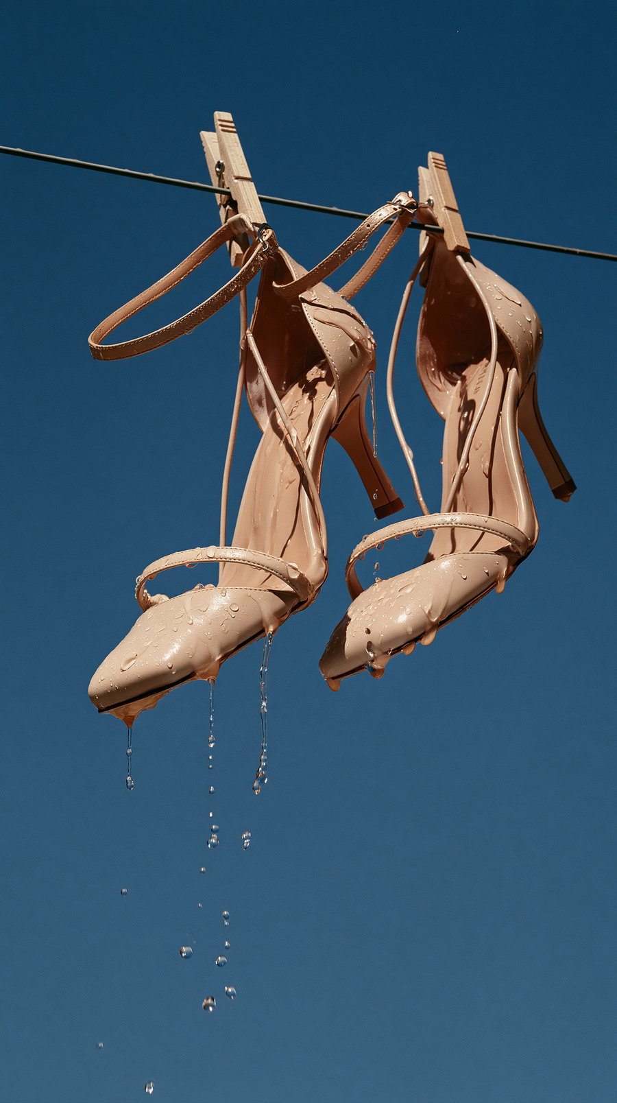 pair of beige high heel shoes wet and hanging on a wire with clothespins against a clear blue sky