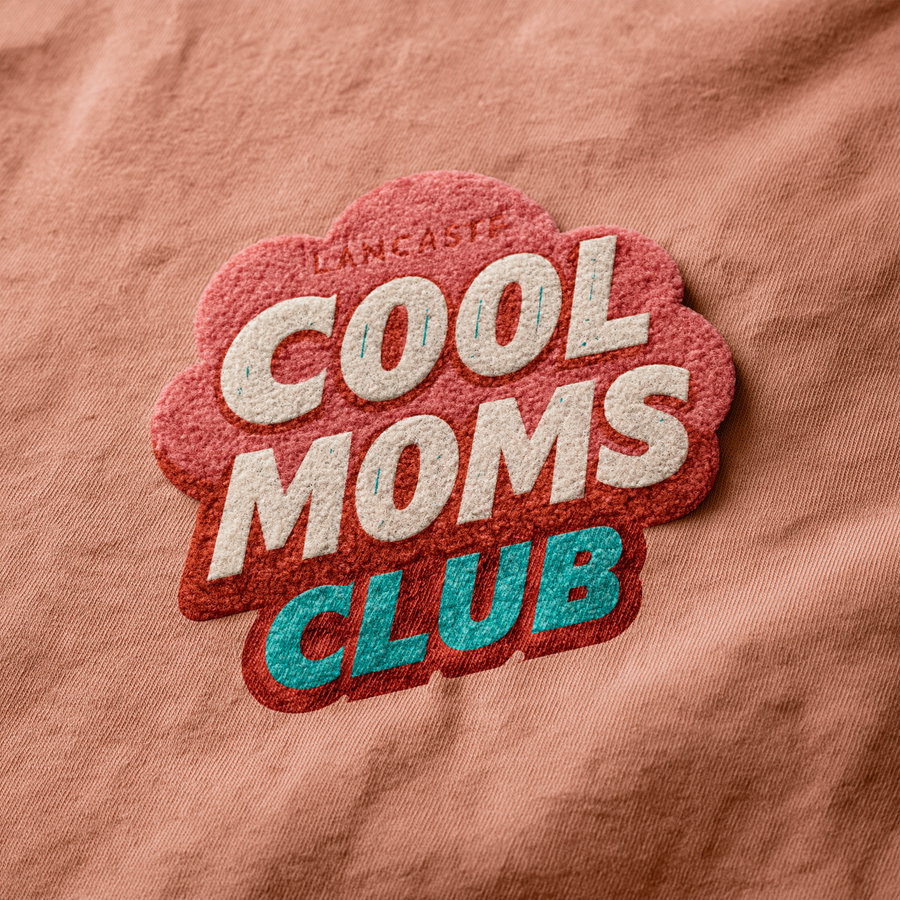 A close-up image of a retro style graphic design printed on fabric with the words Cool Moms Club in cream, red, and teal colors on a pink background