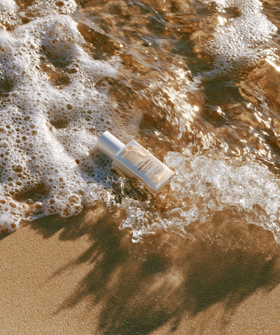 A white skincare bottle lying on wet sand with ocean foamy waves splashing around it in sunlight