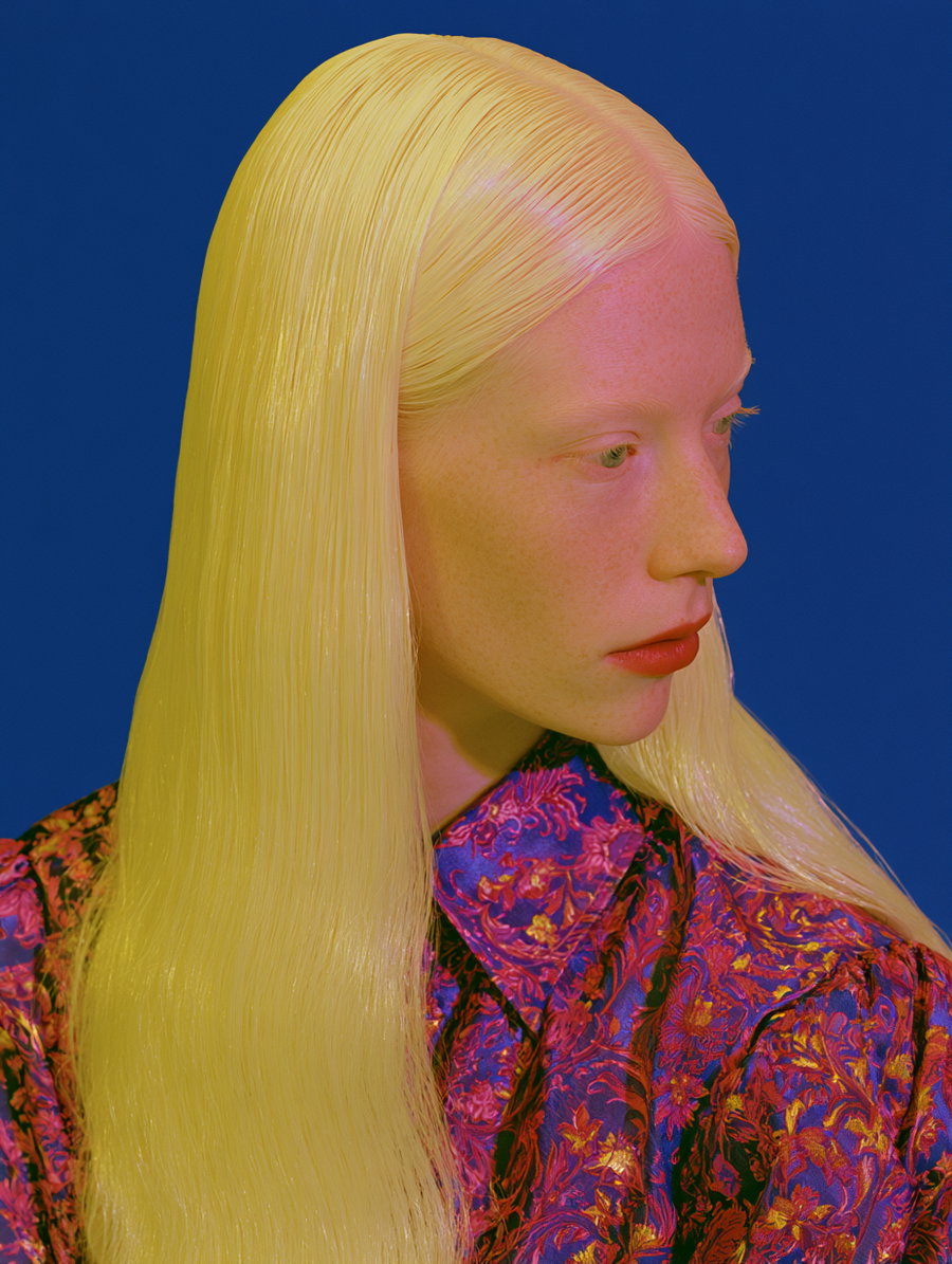 Close-up portrait of a woman with long straight blonde hair, pale skin with freckles, red lips, and wearing a vibrant floral patterned shirt against a solid blue background