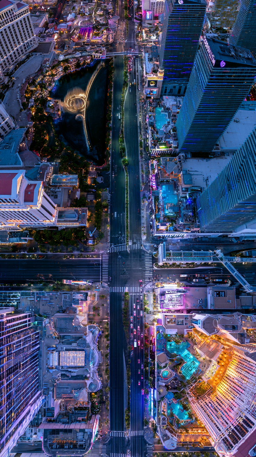 High-angle aerial photo of a city intersection with illuminated buildings, streets with vehicles, a water fountain and pools at night