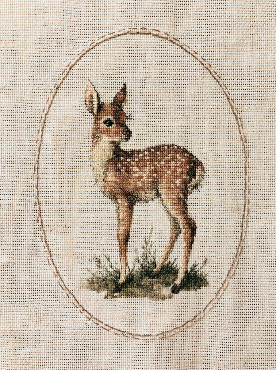 Handcrafted embroidery of a small fawn standing on grass with an oval stitched border on beige fabric