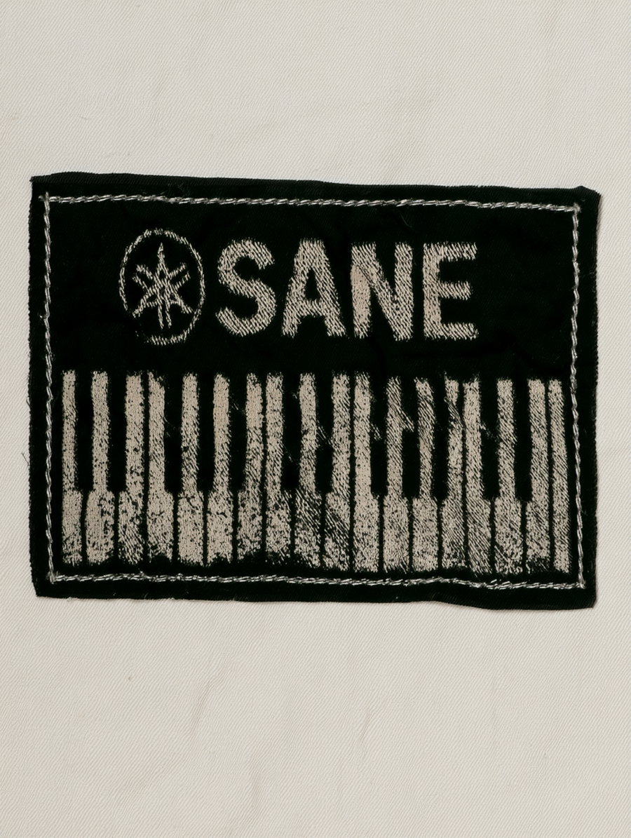 A black fabric patch embroidered with white thread featuring a piano keyboard pattern and the word SANE above it, on a white background.