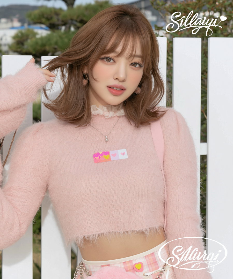 A close-up portrait of a woman wearing a soft pink cropped sweater and light makeup, posing outdoors with one hand touching her hair, standing in front of a white fence.