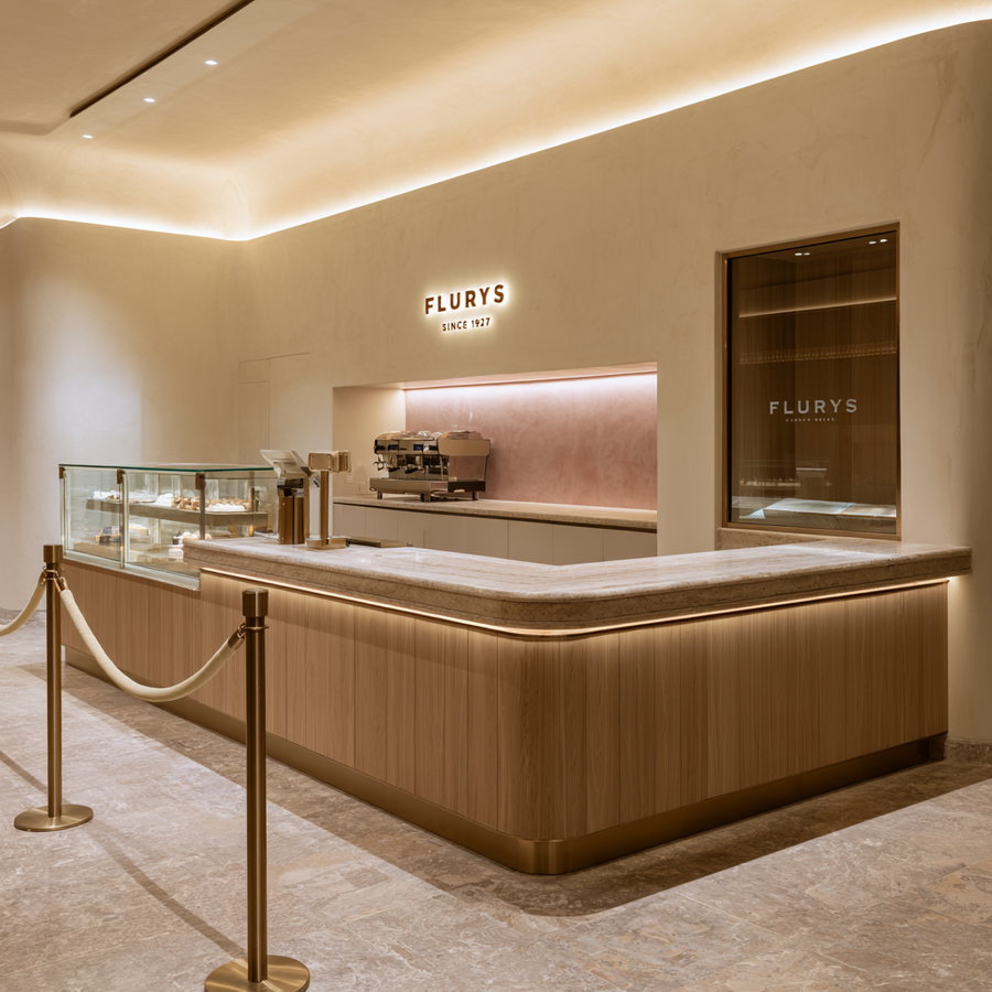Modern coffee shop interior with a curved wooden counter, marble top, gold stanchions, warm wall lighting, and a display case with baked goods