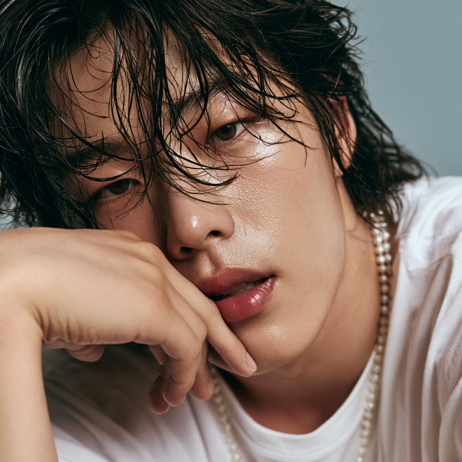 A close-up portrait of a young individual with wet, tousled dark hair partially covering the face, wearing a white shirt and a pearl necklace, resting their chin on their hand against a plain grey background.