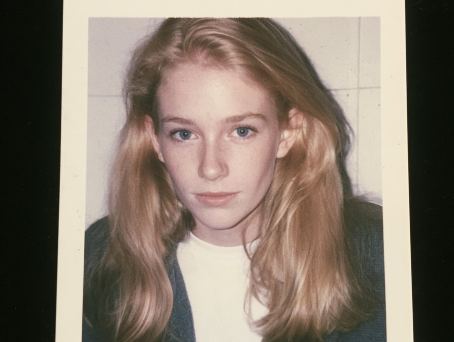 A vintage photographic portrait of a young woman with long blonde hair, blue eyes, wearing a white shirt and dark cardigan, looking directly at the camera against a light tiled background.