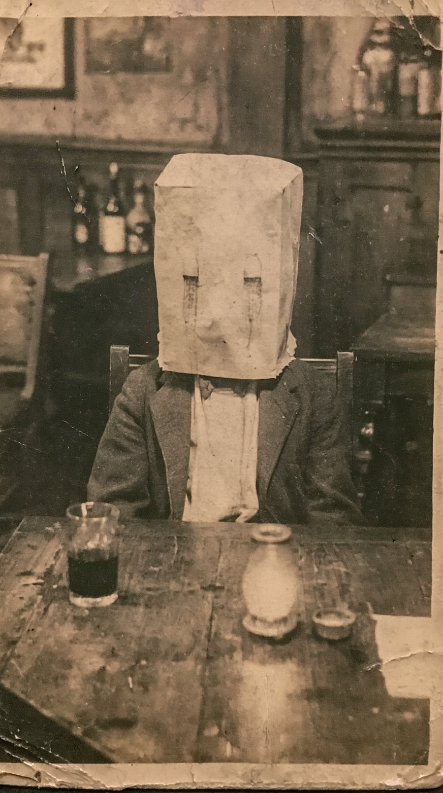 Black and white vintage photo showing a person wearing a suit and a paper bag covering their head sitting at a wooden table with a glass and pottery items on it