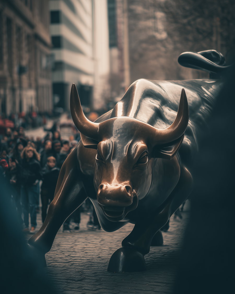 A bronze sculpture of a charging bull on a cobblestone street with blurred crowd and tall buildings in the background