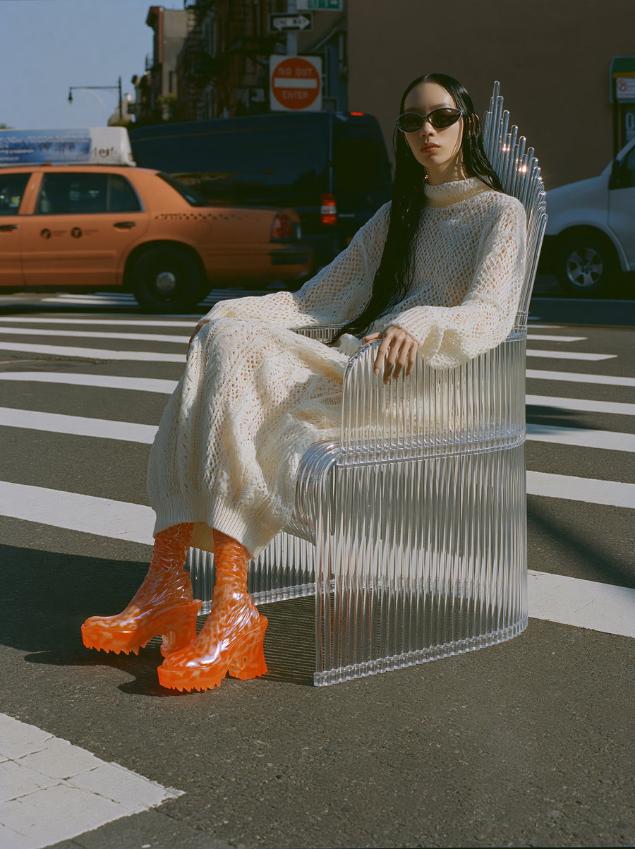 A woman wearing a white knitted dress and orange high-heeled platform boots sits on a transparent modern chair at a pedestrian crosswalk, with urban background including cars and street signs.