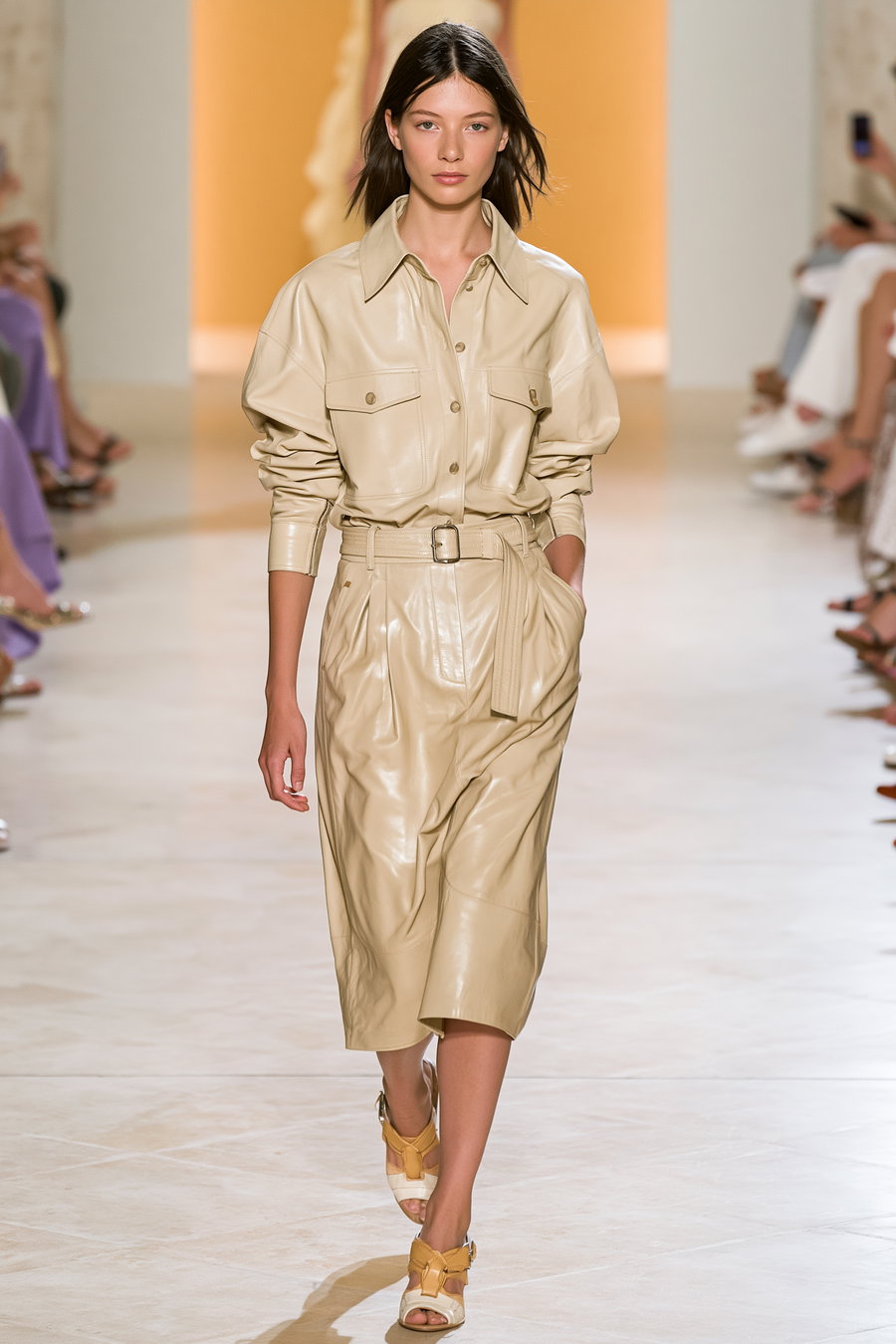 A female model walking on a fashion runway wearing a beige leather belted shirt and midi skirt with matching yellow and white heels