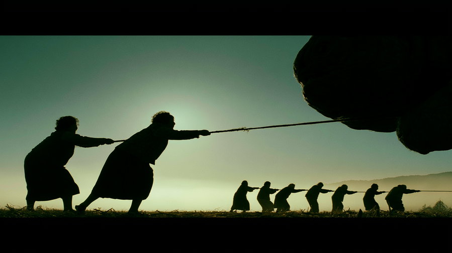 Silhouettes of people pulling a large rope against a greenish sky with the sun behind them, outdoors on grassy land