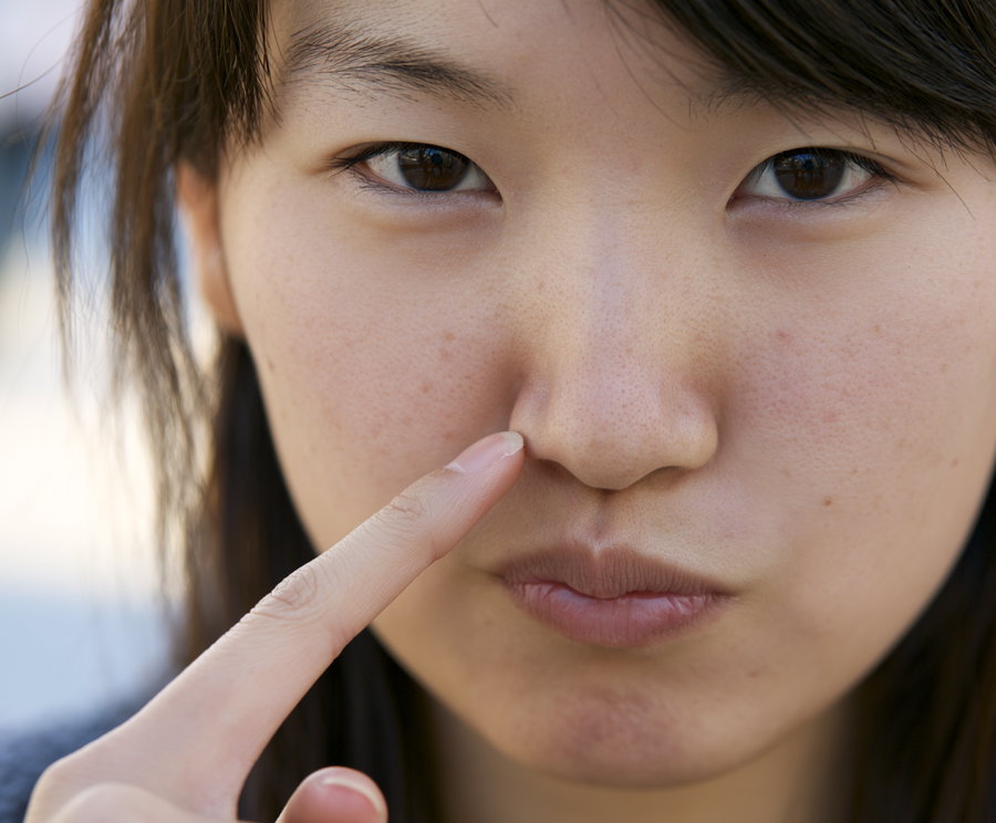 Close-up of a young woman with brown eyes and natural skin, touching the side of her nose with her index finger.