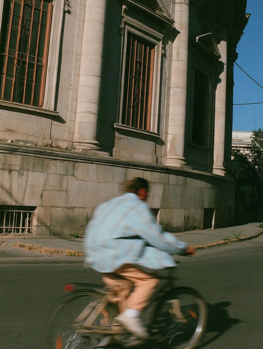 A blurred person wearing light-colored clothing rides a bicycle on a street next to a stone building with large windows and columns under a clear blue sky