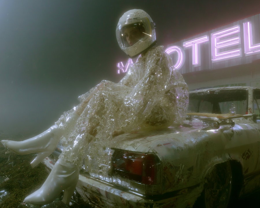 Person wearing a translucent sequined outfit and white boots with a clear helmet sitting on the hood of a worn vintage car under a neon motel sign in foggy atmosphere