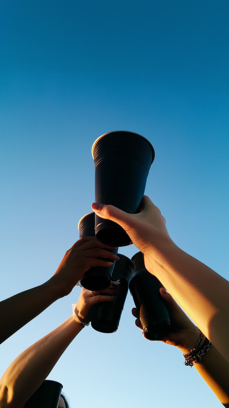 Several hands holding black plastic cups raised together with a clear blue sky background