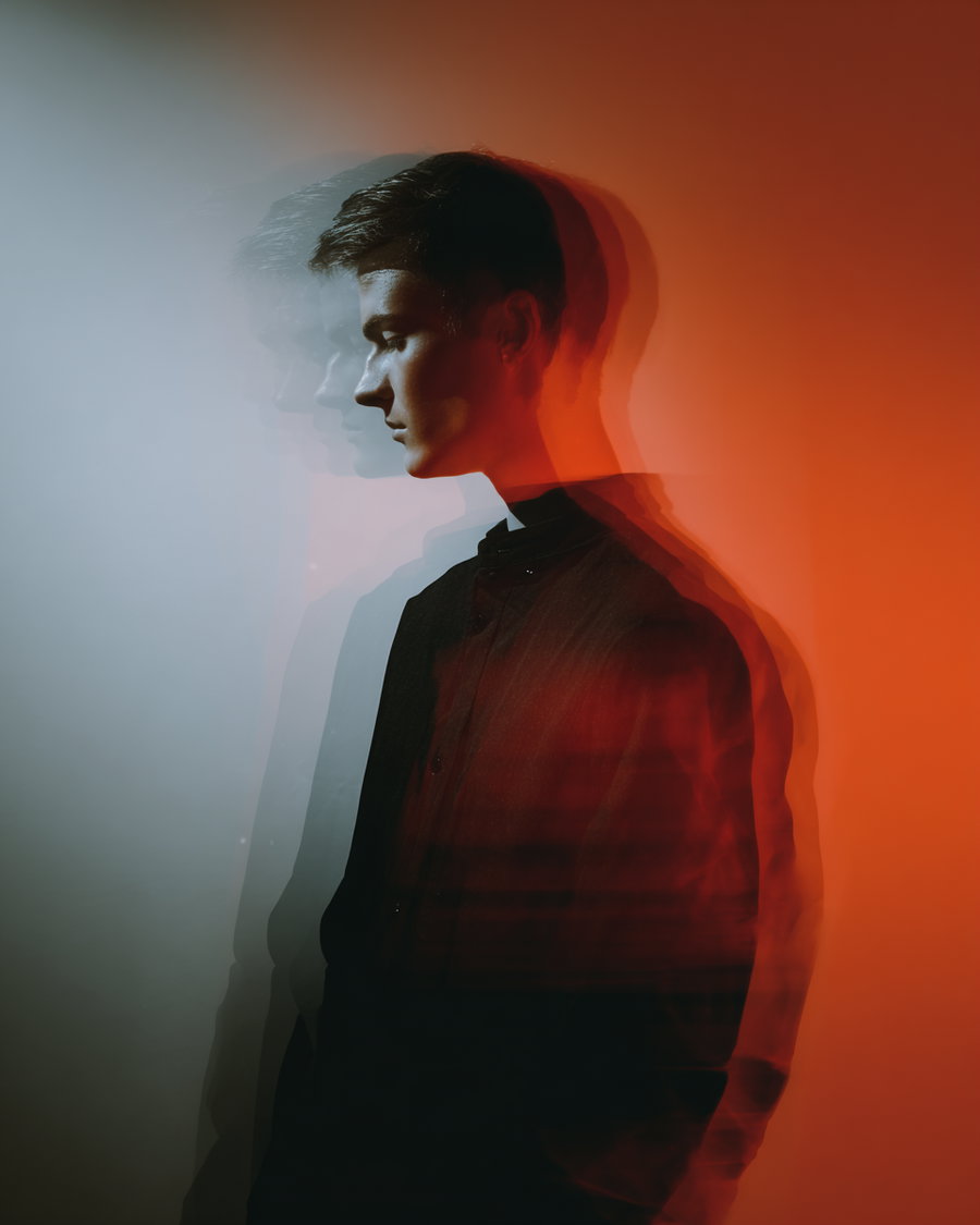 side view of a young man in dark clothing with a double exposure or motion blur effect, against a background split between cool blue and warm red lighting