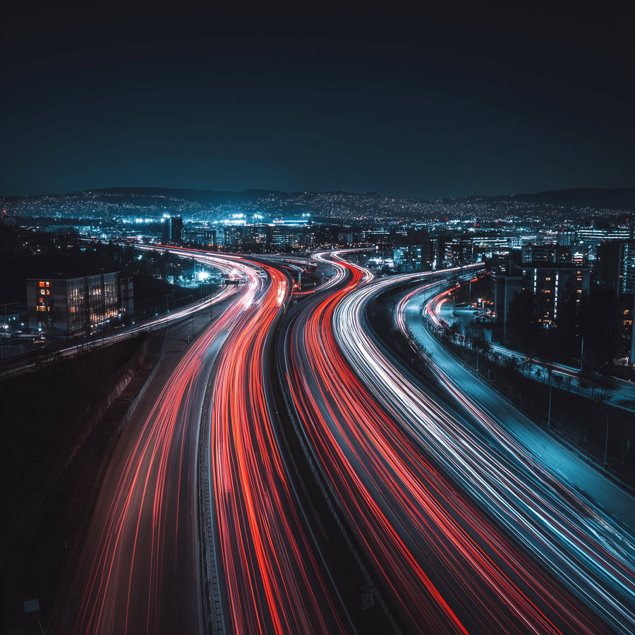 a night cityscape showing busy highways with streaks of red and white lights from moving vehicles, surrounded by city buildings and distant hills under a dark sky