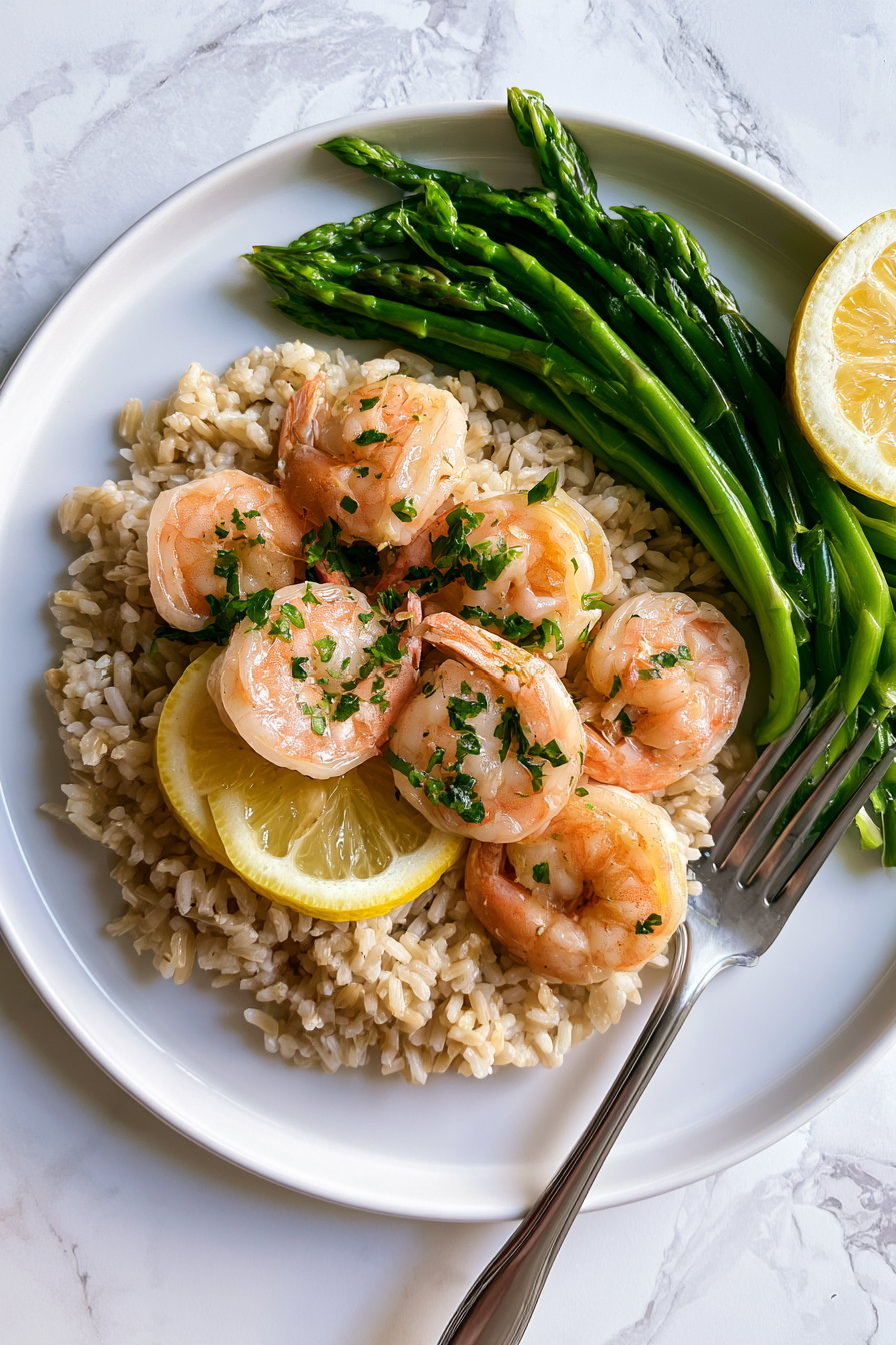 Close-up of a white plate with cooked shrimp garnished with herbs, lemon slices, asparagus, and brown rice with a fork on the side