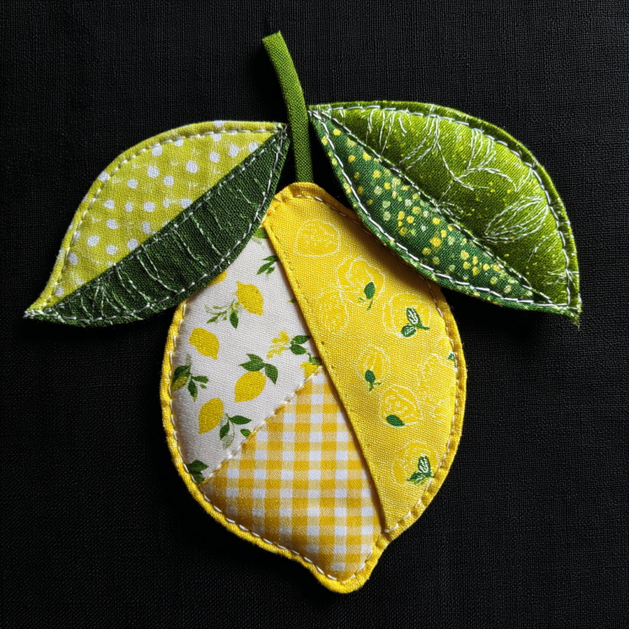 Crafted fabric art patch featuring a lemon shape composed of yellow and patterned fabric pieces with two green fabric leaves on a black background