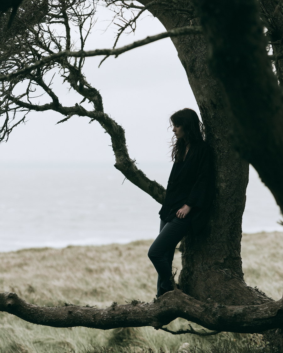A woman in dark clothing leaning against a tree with bare branches, standing on grass with a lake in the background under an overcast sky
