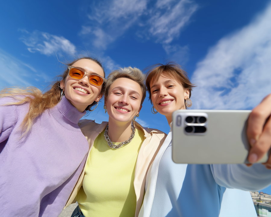 Three young women smiling closely together, one holding a smartphone to take a selfie, against a backdrop of partly cloudy blue sky