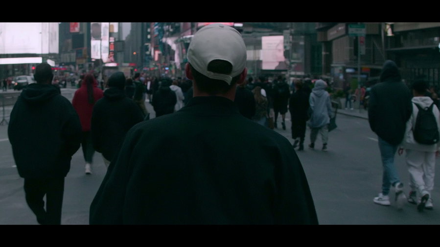 A person wearing a white cap and dark jacket is walking among a crowd of people on a city street with buildings and billboards visible in the background.