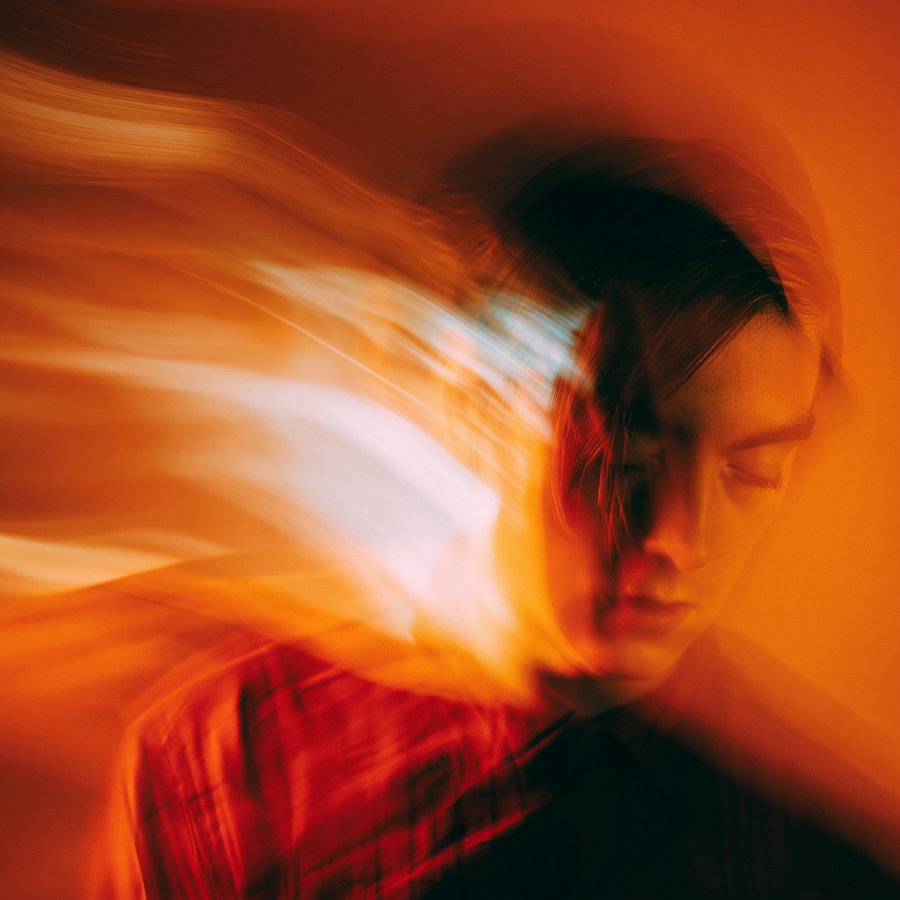 A blurred and abstract portrait of a person with eyes closed, surrounded by dynamic orange and red light streaks creating a moody effect