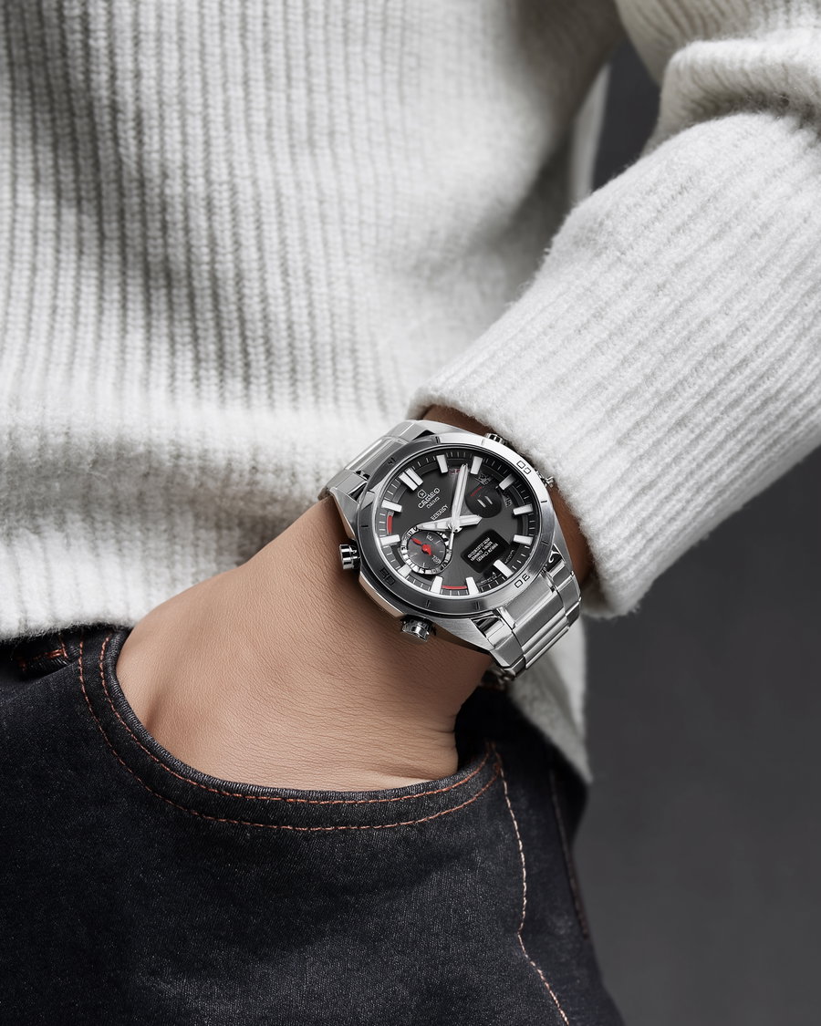 A close-up photo of a person's wrist wearing a modern silver metal strap watch with a black dial, white hour markers, and red accent marks, partially inside a dark denim pocket and a white ribbed sweater sleeve