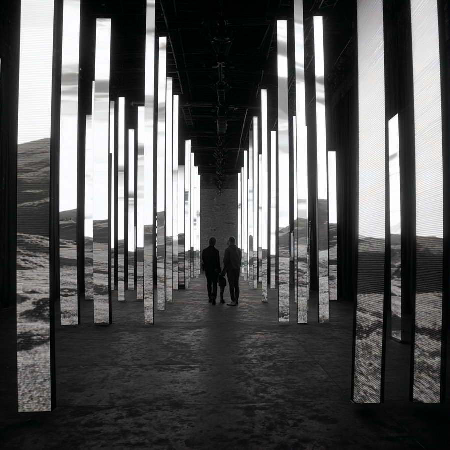 Black and white photo of an art installation featuring tall, vertical, illuminated rectangular panels arranged in rows with two people walking in the center