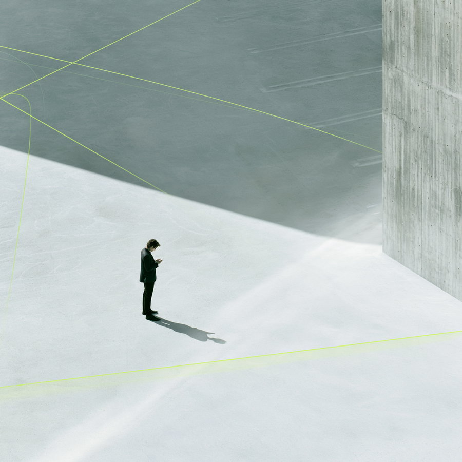 A man in a black suit stands alone on a concrete surface with distinct sharp shadows and light beams, next to a tall concrete wall